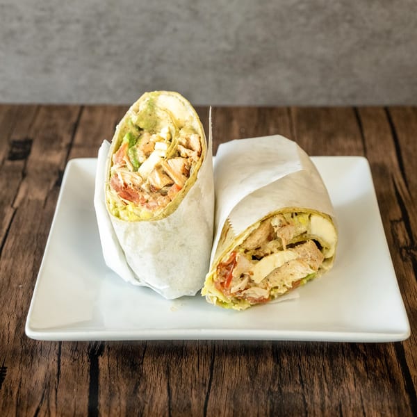 Two chicken wraps with avocado, tomato, and lettuce, one cut in half, on a white plate.