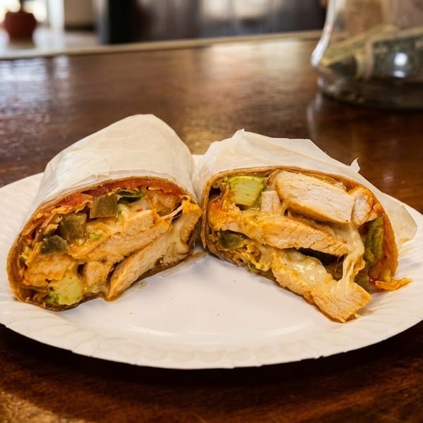 A chicken wrap, cut in half, on a white paper plate, filled with chicken, avocado, and cheese.