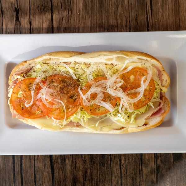 A large deli sub sandwich with ham, cheese, lettuce, tomatoes, and onions on a white plate.