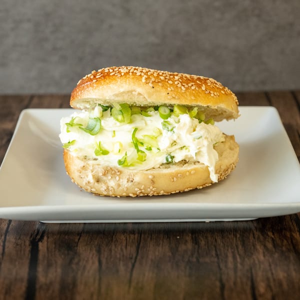 Sesame bagel with a generous spread of cream cheese and chopped green onions on a white plate.