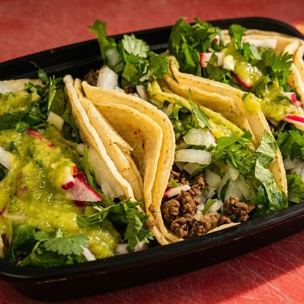 Four tacos in a black container, filled with meat, onions, cilantro, radishes, and green salsa.